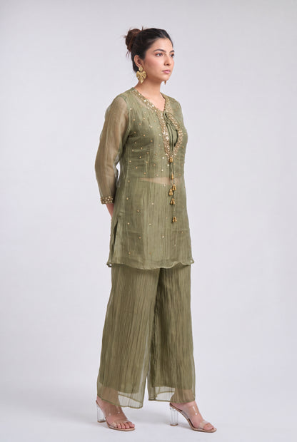 A woman modeling a sage green three-piece ethnic suit with gold embroidery, featuring a long-sleeve tunic, pleated wide-leg pants, and a matching dupatta, set against a plain light gray background.
