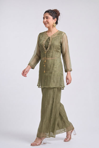 A woman modeling a three-piece olive green ethnic suit with sheer long sleeves, a tie-neck embroidered tunic, and matching pleated pants, photographed against a light gray studio background.