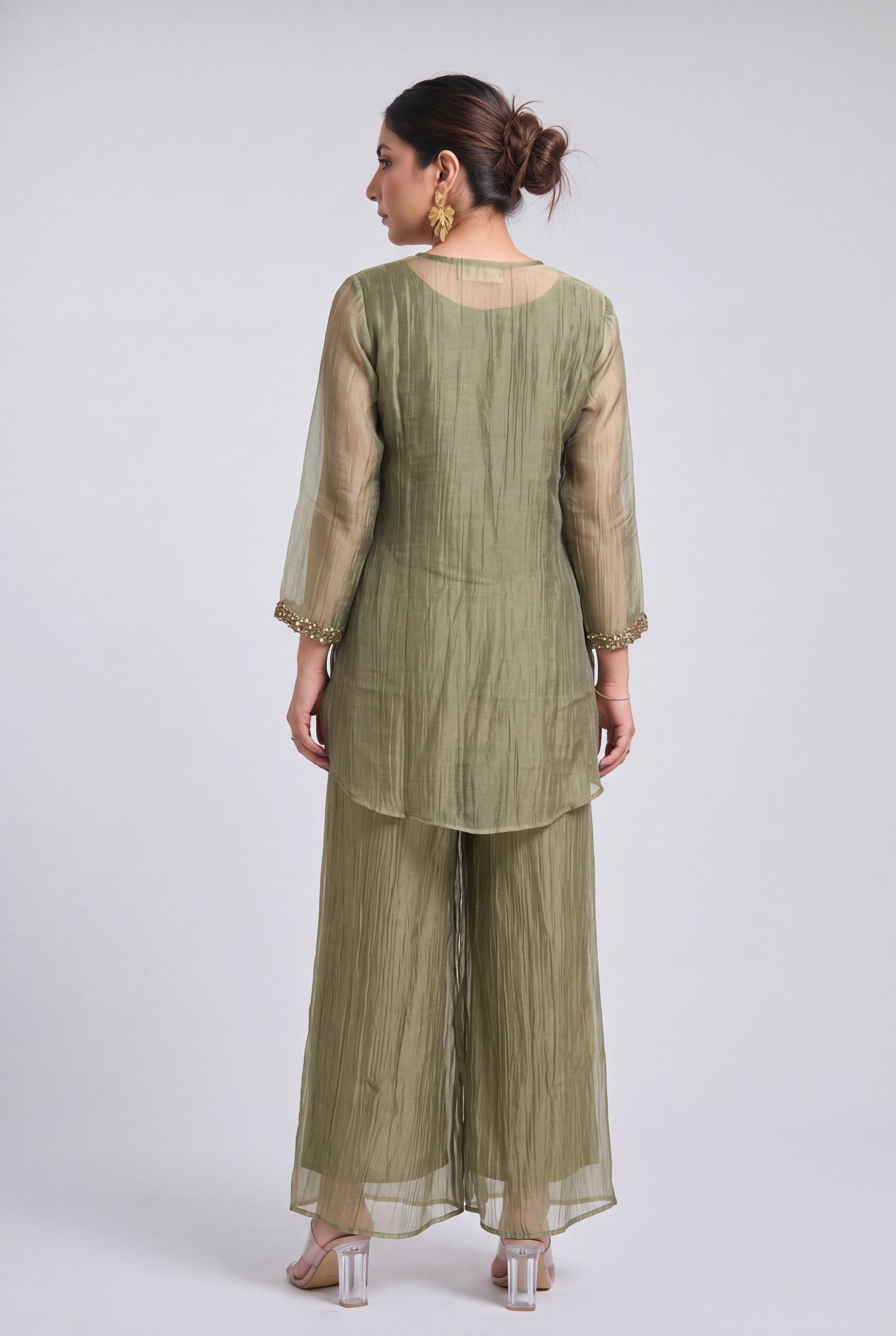 A woman shown from the back wearing an olive green traditional outfit with sheer sleeves and wide-leg pants, photographed against a light gray studio background.