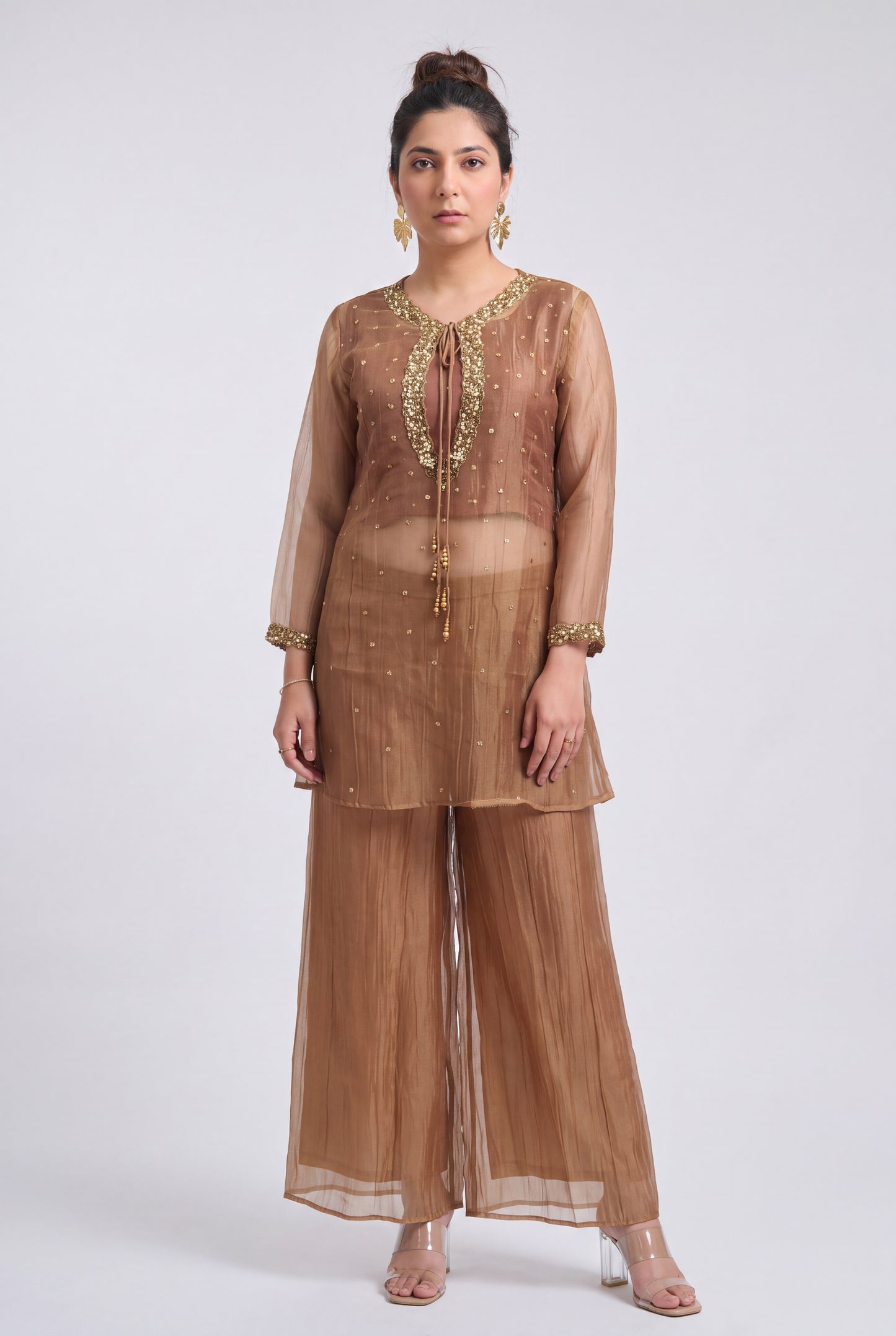 A woman models a three-piece sheer brown organza ethnic suit with gold embroidery, featuring a long-sleeve kurta, wide-leg pants, and a matching dupatta, set against a plain light gray background.