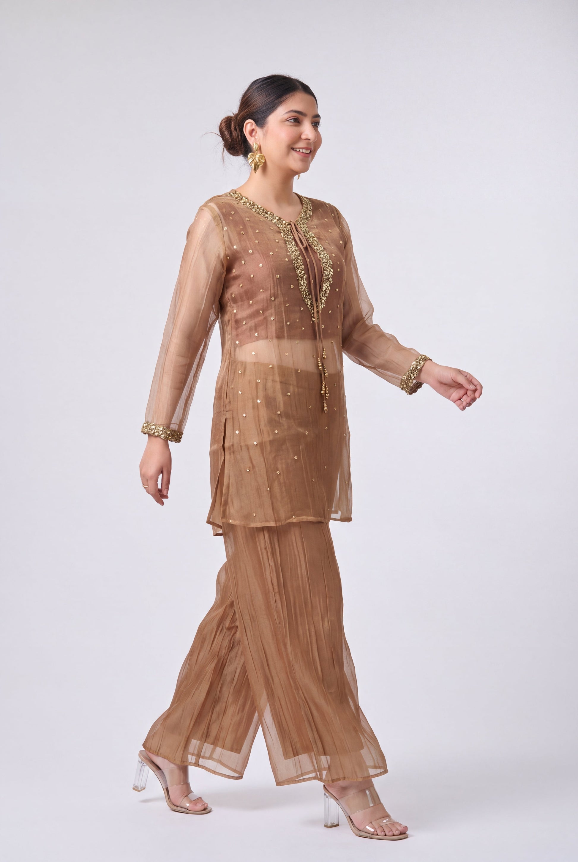 A woman models a three-piece beige organza ethnic suit with gold embroidery, featuring a long-sleeve tunic, pleated pants, and a matching dupatta, set against a plain light gray background.