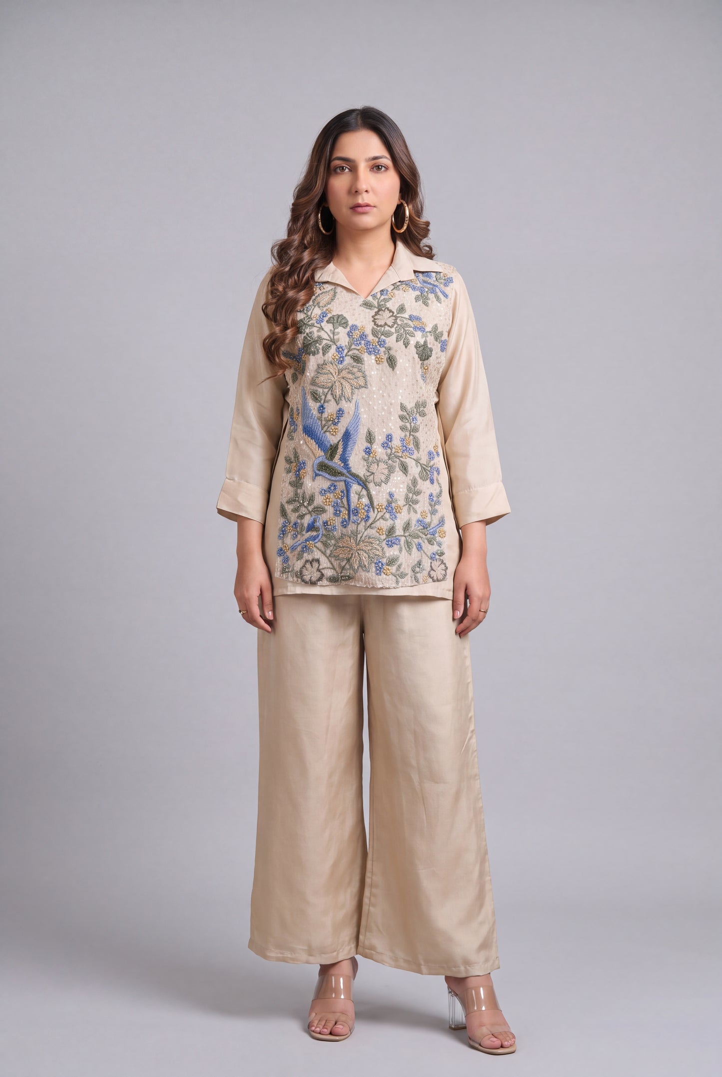 A woman modeling a beige two-piece outfit consisting of a long-sleeve collared tunic top with blue and green floral embroidery and matching wide-leg trousers, shot against a solid gray studio background.