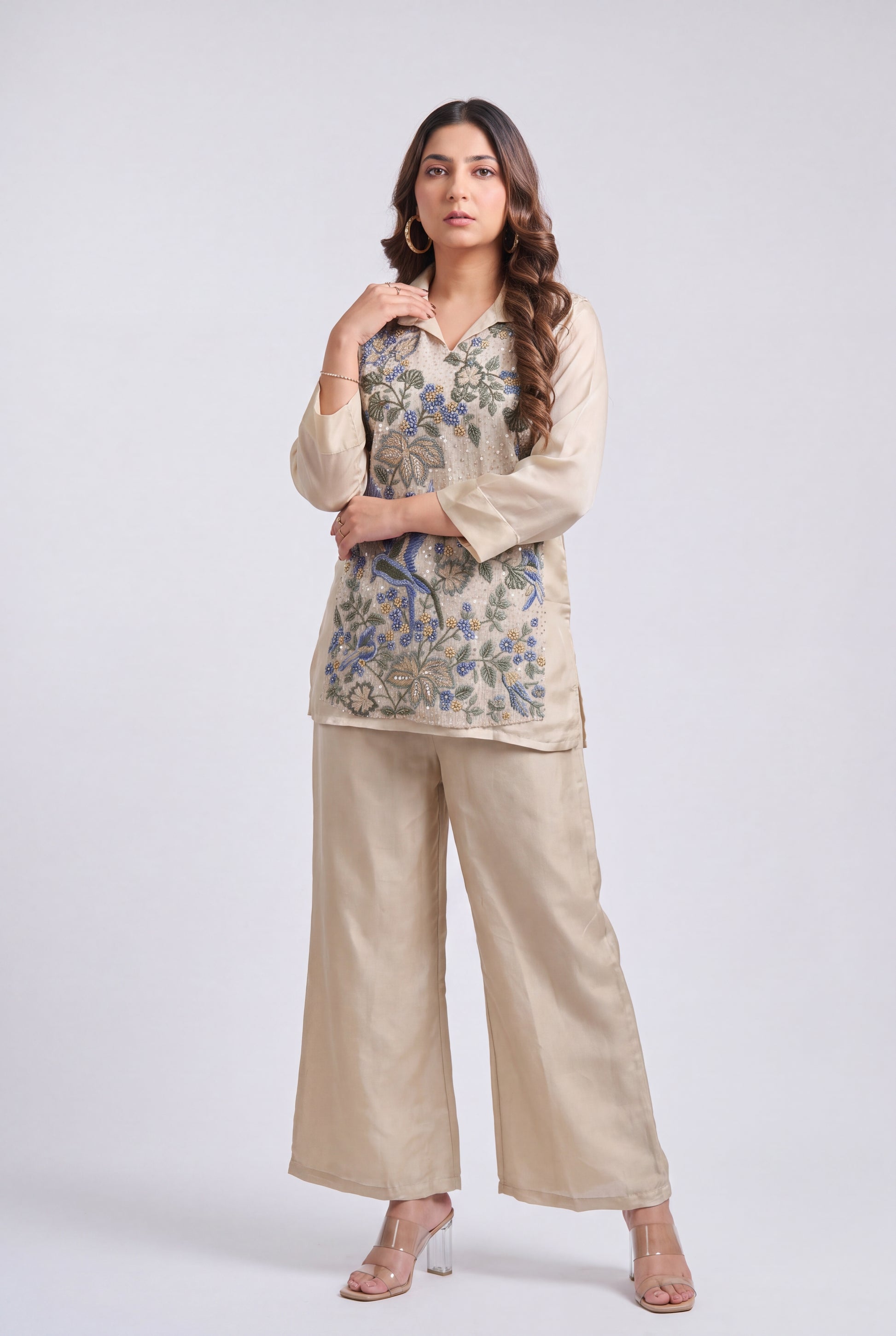 A woman modeling a beige two-piece kurta set with a blue and green floral embroidered tunic top and matching wide-leg trousers, photographed against a light gray studio background.