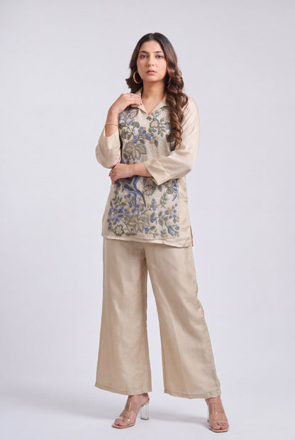 A woman modeling a beige two-piece kurta set with a blue and green floral embroidered tunic top and matching wide-leg trousers, photographed against a light gray studio background.