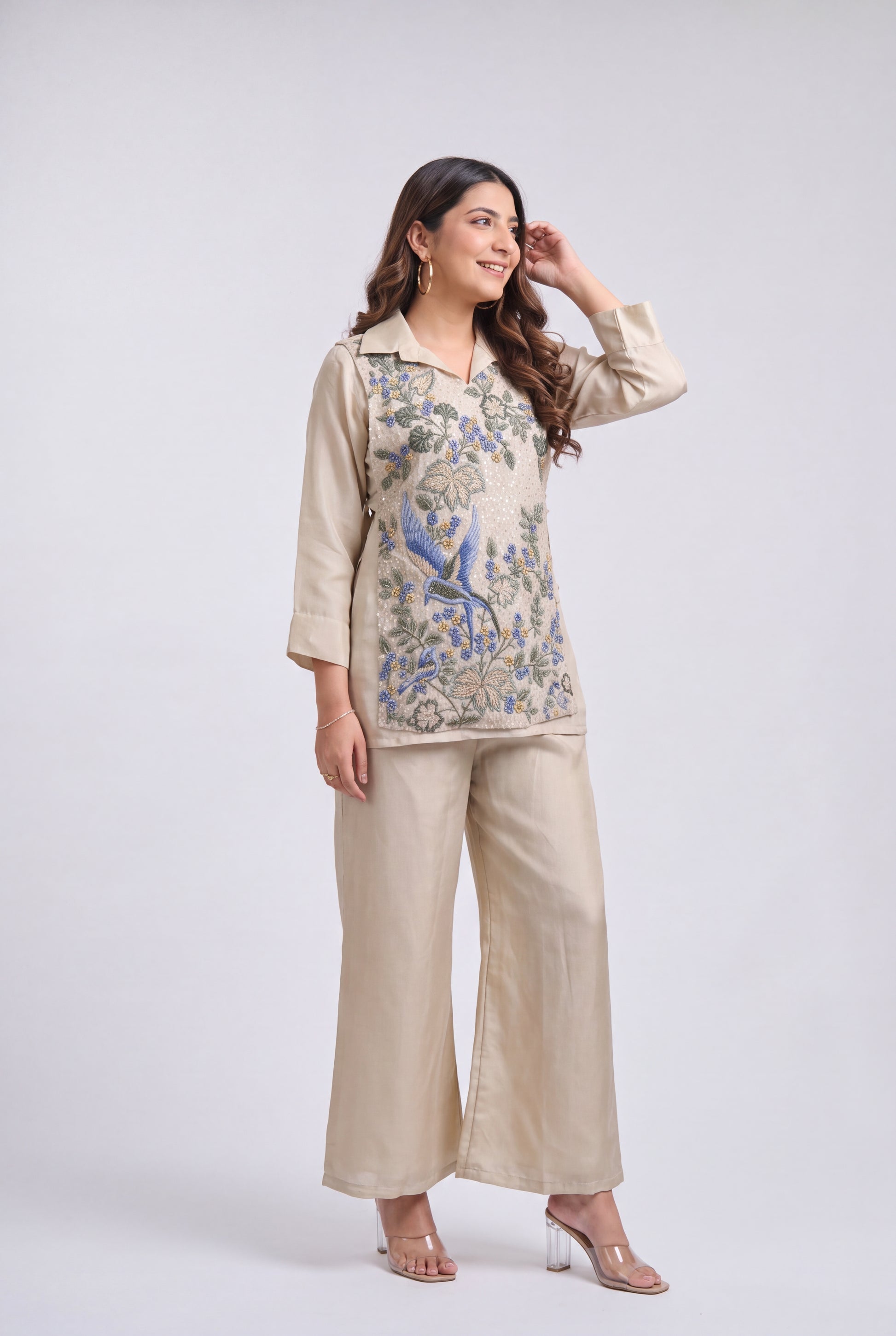 A woman modeling a beige two-piece outfit consisting of a long-sleeve top with blue and green floral embroidery and matching wide-leg trousers, set against a plain light gray studio background.