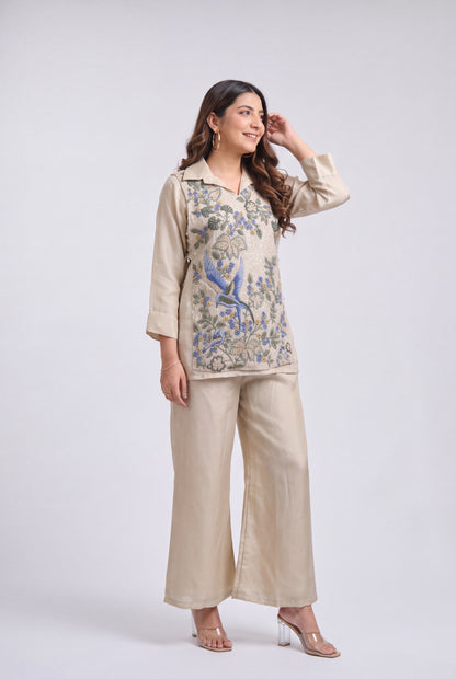 A woman modeling a beige two-piece outfit consisting of a long-sleeve top with blue and green floral embroidery and matching wide-leg trousers, set against a plain light gray studio background.