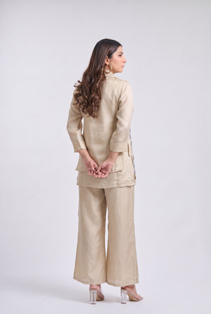 A woman is shown from the back wearing a light beige two-piece outfit consisting of a long-sleeve jacket and wide-leg pants, paired with clear high-heeled sandals, against a light gray studio background.