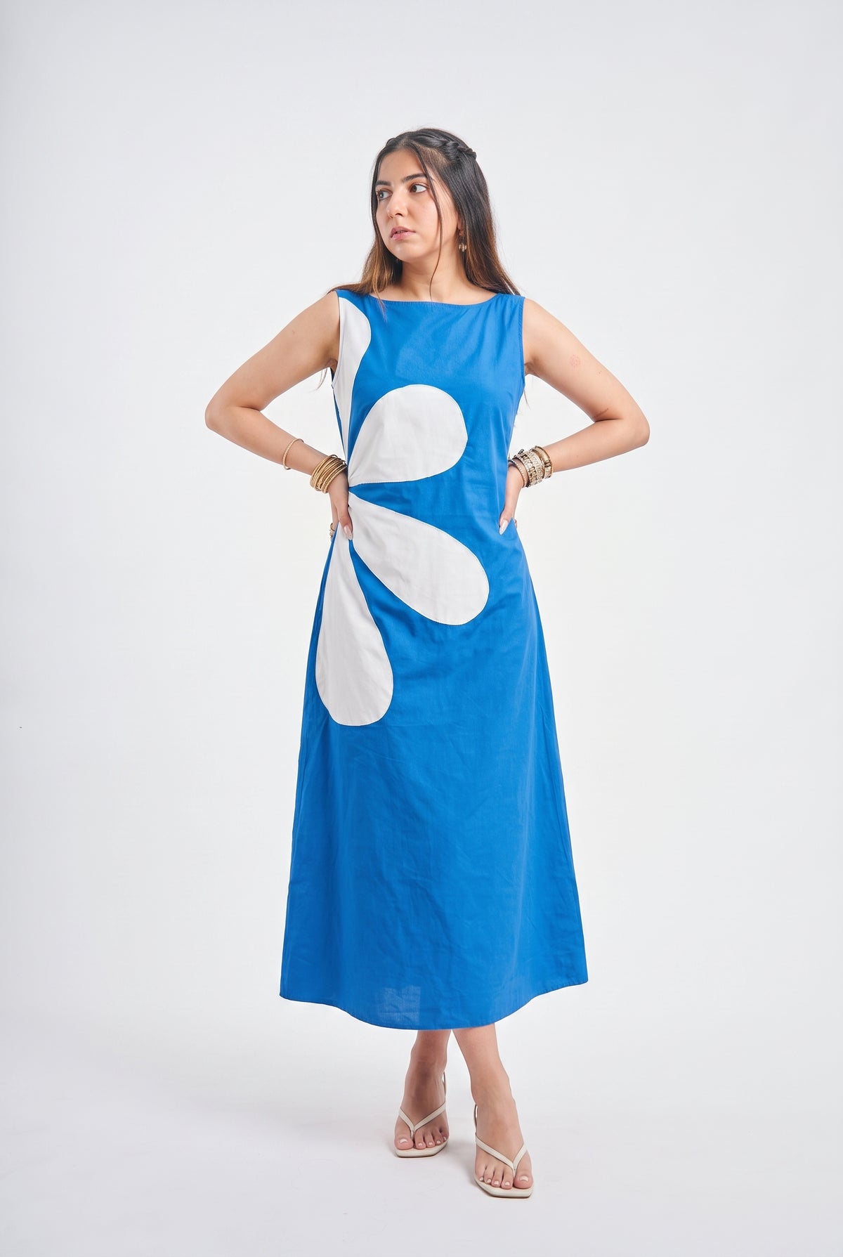 A woman modeling a sleeveless blue midi dress with a large white abstract petal-like graphic on the front, posed against a light gray studio background.