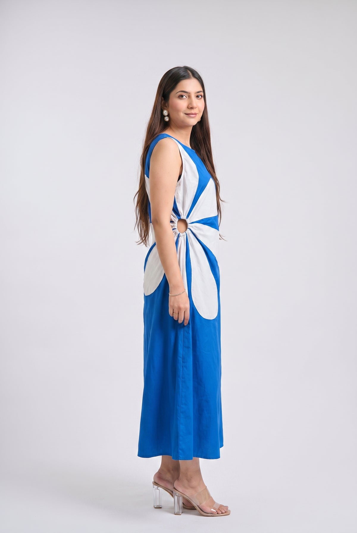 A woman modeling a sleeveless blue maxi dress with a large white abstract floral or ribbon-like panel across the back and waist, paired with clear high-heel sandals, against a light gray studio background.