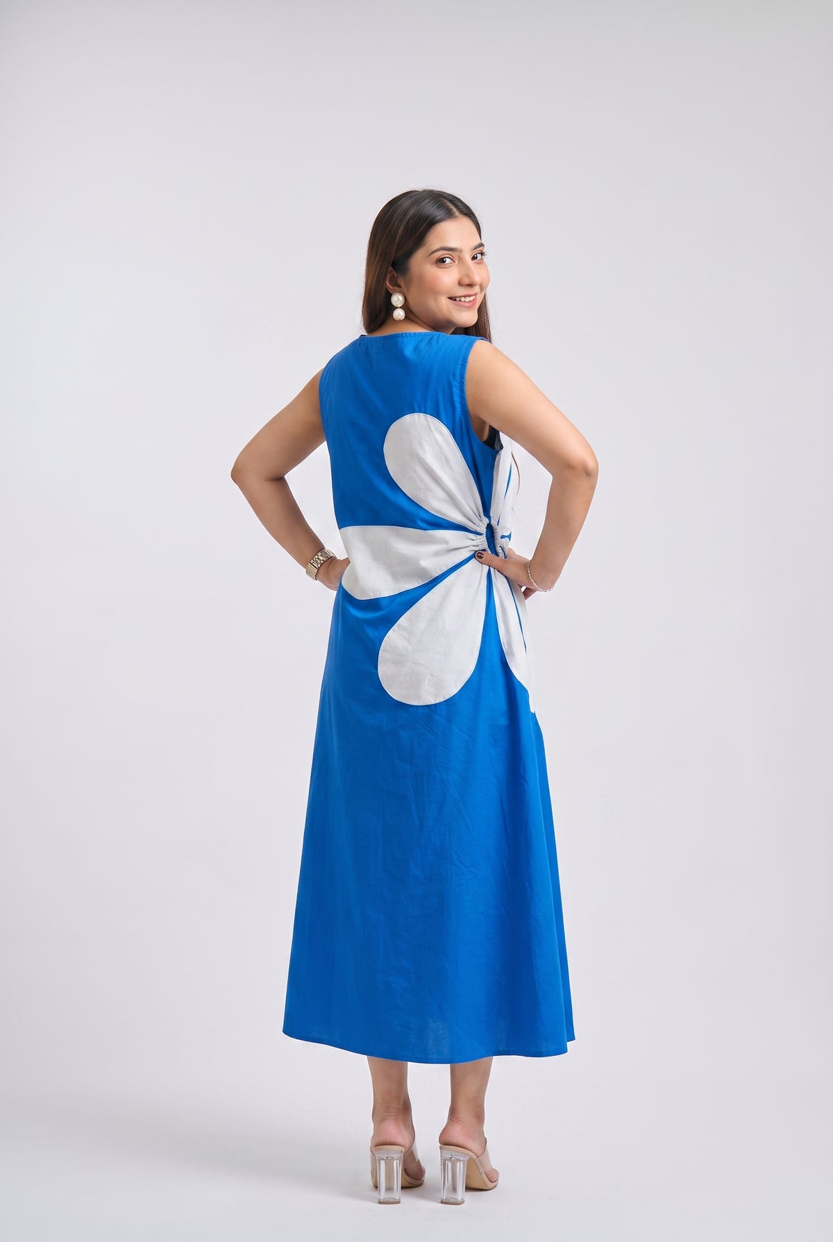 A woman modeling a sleeveless royal blue maxi dress from the back, featuring a large white abstract shape on the back panel, posed against a light gray studio background.