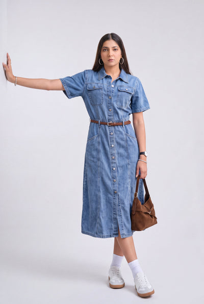 A woman modeling a short-sleeve blue denim midi dress with a button-down front, chest pockets, and a brown belt. She is wearing white socks, white sneakers, and holding a brown leather shoulder bag against a plain light gray studio background.