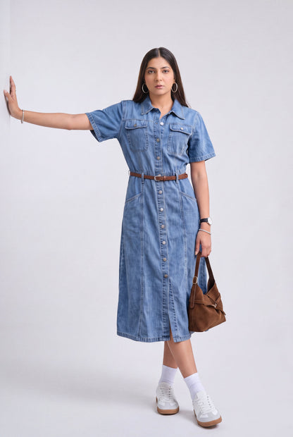 A woman modeling a short-sleeve blue denim midi dress with a button-down front, chest pockets, and a brown belt. She is wearing white socks, white sneakers, and holding a brown leather shoulder bag against a plain light gray studio background.