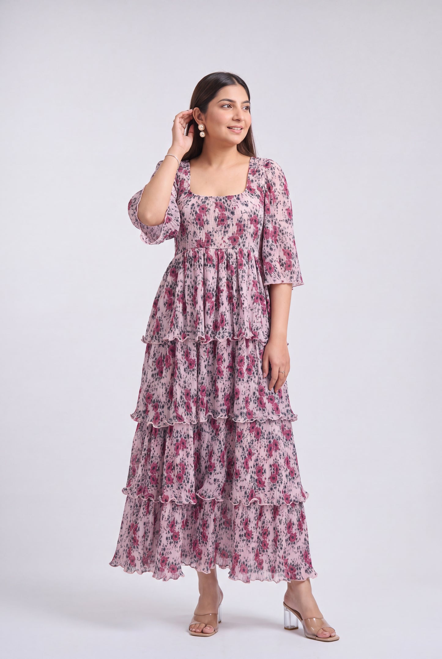 A woman shown from the wearing a tiered maxi dress with a pink and purple floral print on a white base, photographed against a light gray studio background.