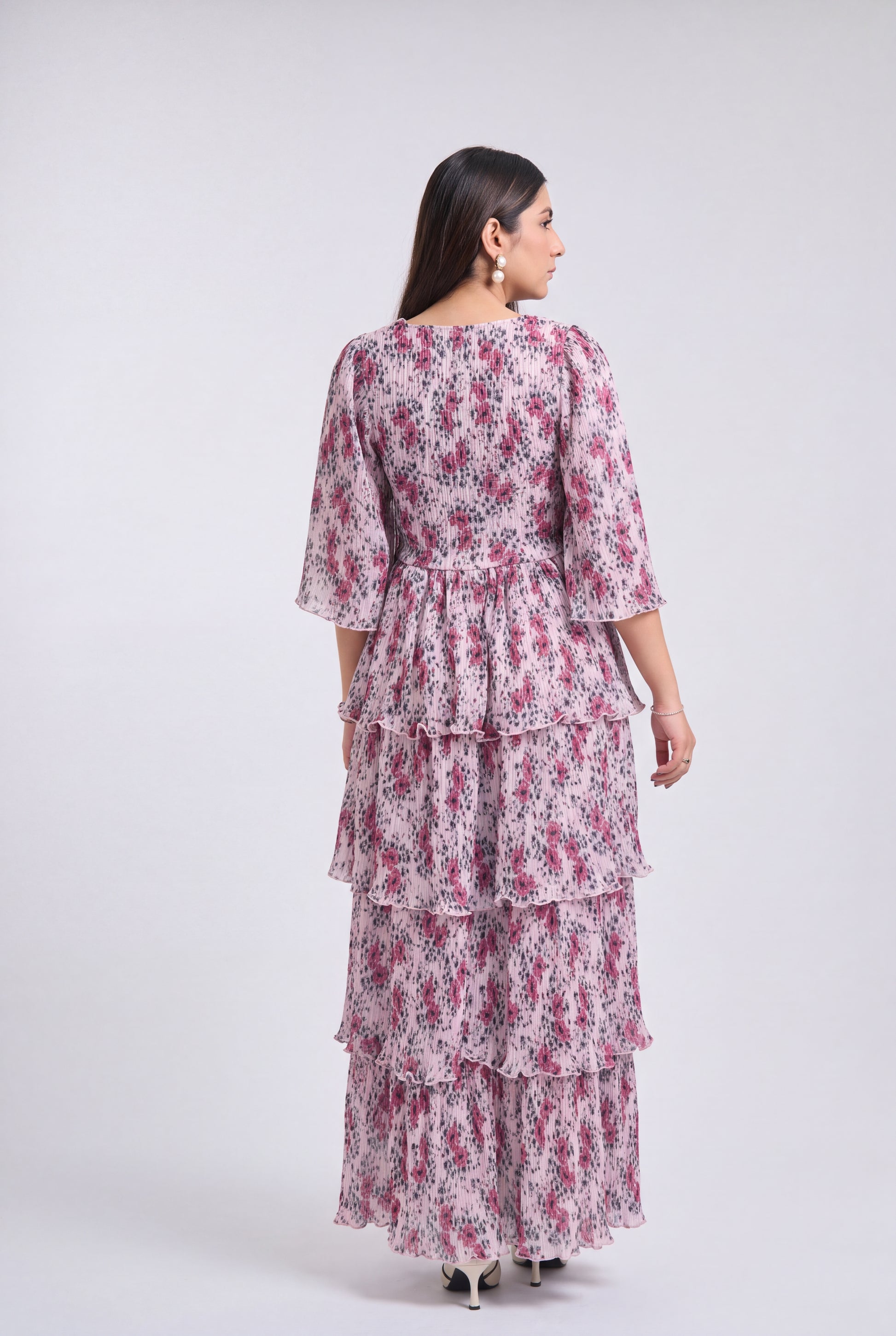 A woman shown from the back wearing a tiered maxi dress with a pink and purple floral print on a white base, photographed against a light gray studio background.
