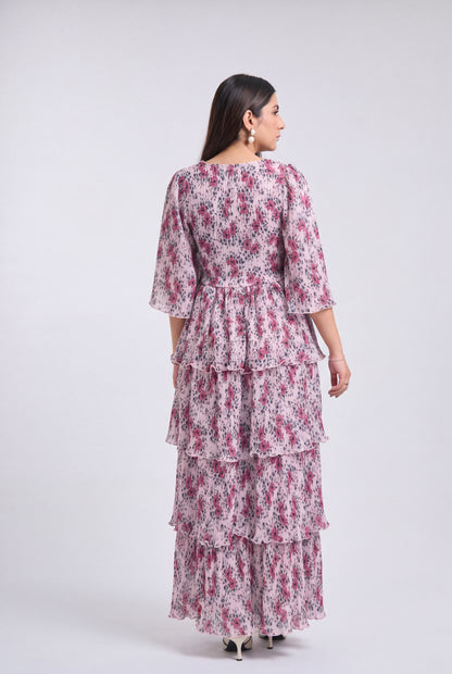 A woman shown from the back wearing a tiered maxi dress with a pink and purple floral print on a white base, photographed against a light gray studio background.