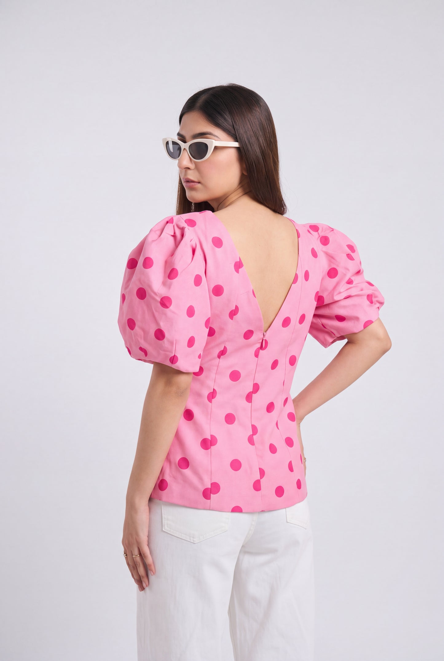 A woman shown from the back wearing a pink puff-sleeve top with dark pink polka dots and a deep V-neckline, paired with white pants against a light gray studio background.