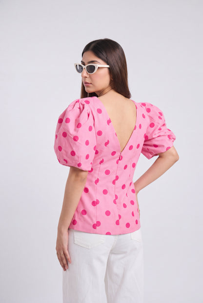 A woman shown from the back wearing a pink puff-sleeve top with dark pink polka dots and a deep V-neckline, paired with white pants against a light gray studio background.