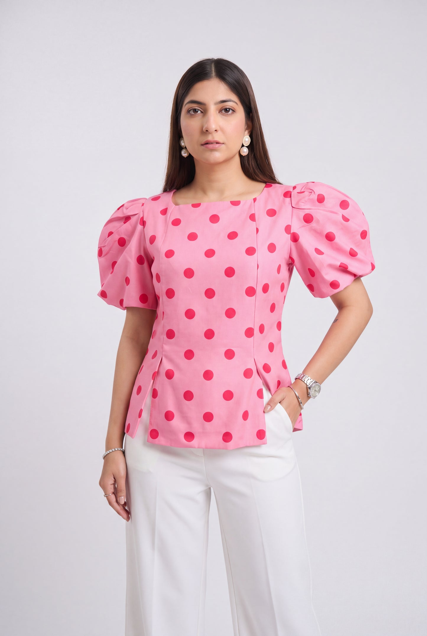 A woman modeling a pink puff-sleeve blouse with red polka dots, paired with white trousers, against a light gray studio background.