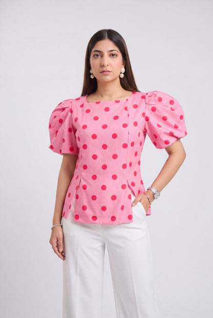 A woman modeling a pink puff-sleeve blouse with red polka dots, paired with white trousers, against a light gray studio background.