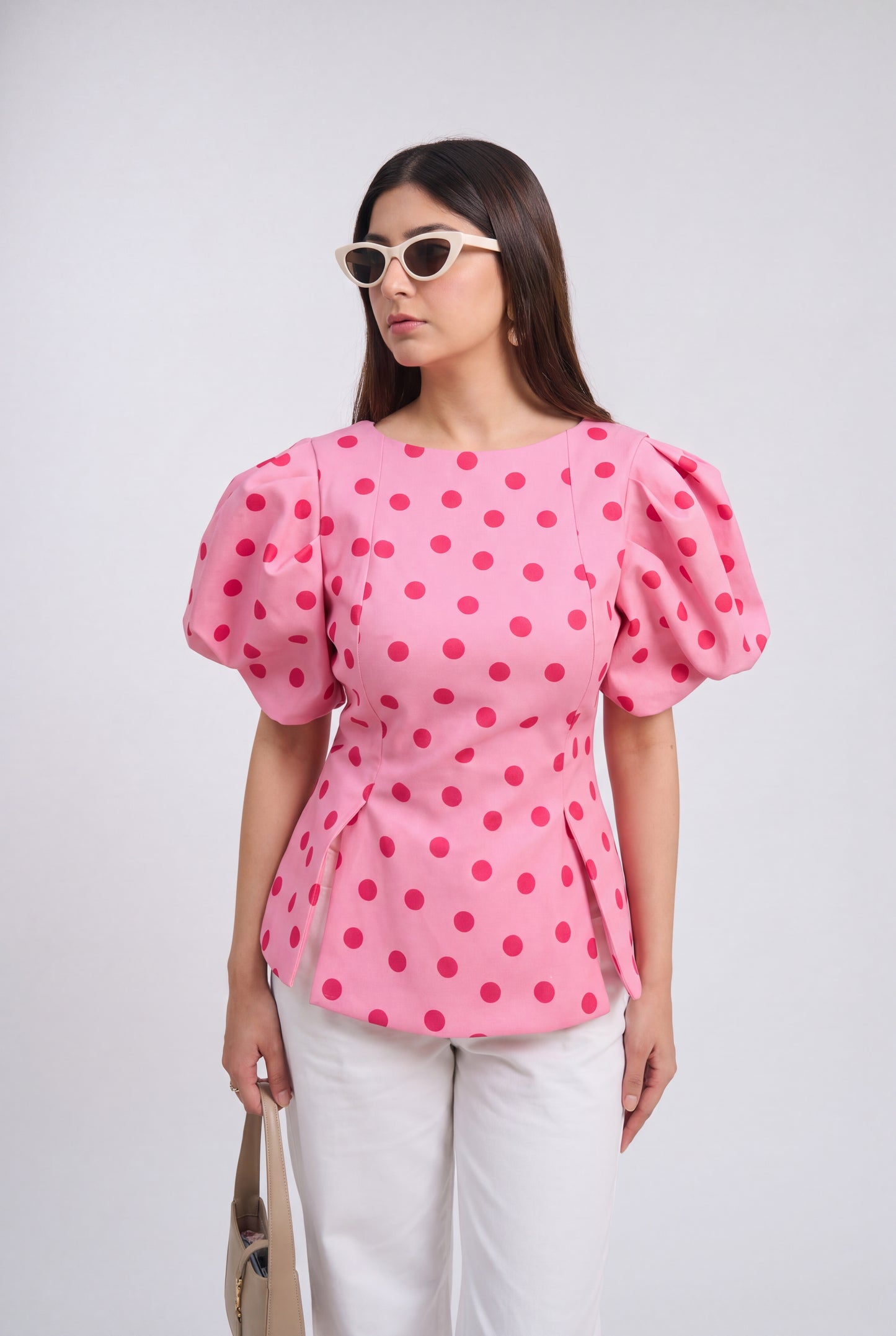 A woman modeling a pink puff-sleeve blouse with a bold magenta polka dot pattern, paired with white pants and white-framed sunglasses, against a light gray studio background.