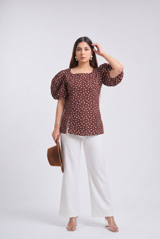 A woman modeling a brown puff-sleeve top with a white polka dot pattern, paired with white wide-leg pants, holding a brown hat against a light gray studio background.