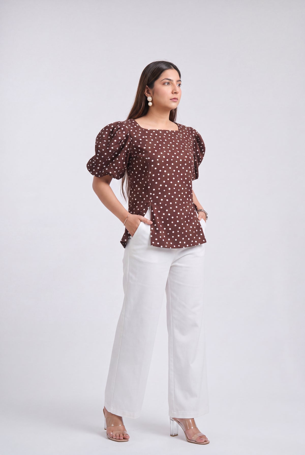 A woman modeling a brown puff-sleeve top with white polka dots, paired with white wide-leg pants and clear high-heeled sandals, against a light gray studio background.