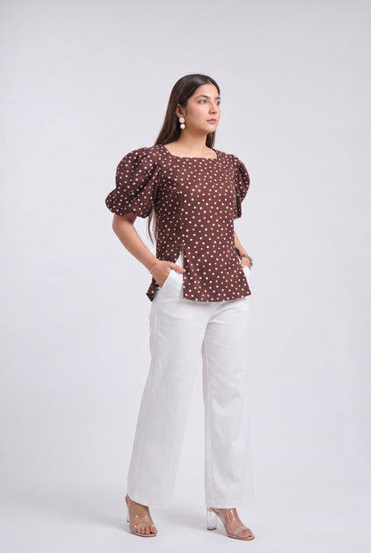 A woman modeling a brown puff-sleeve top with white polka dots, paired with white wide-leg pants and clear high-heeled sandals, against a light gray studio background.