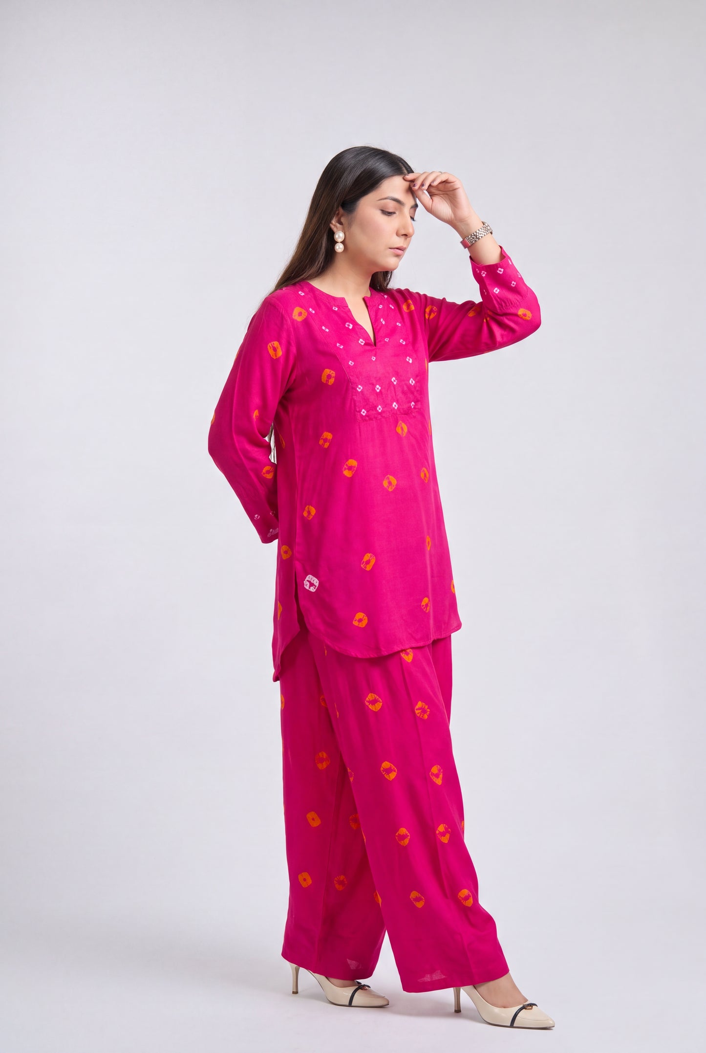 A woman modeling a bright pink two-piece kurta pajama set with orange floral prints, standing against a light gray studio background.
