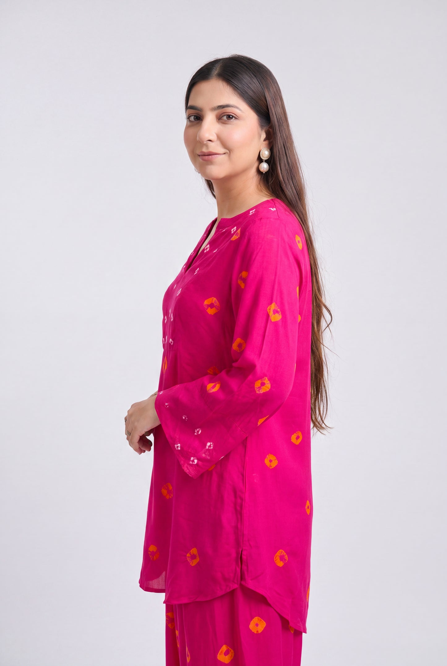 A woman modeling a bright pink traditional kurta set with orange and yellow floral motifs, photographed against a plain light gray studio background.