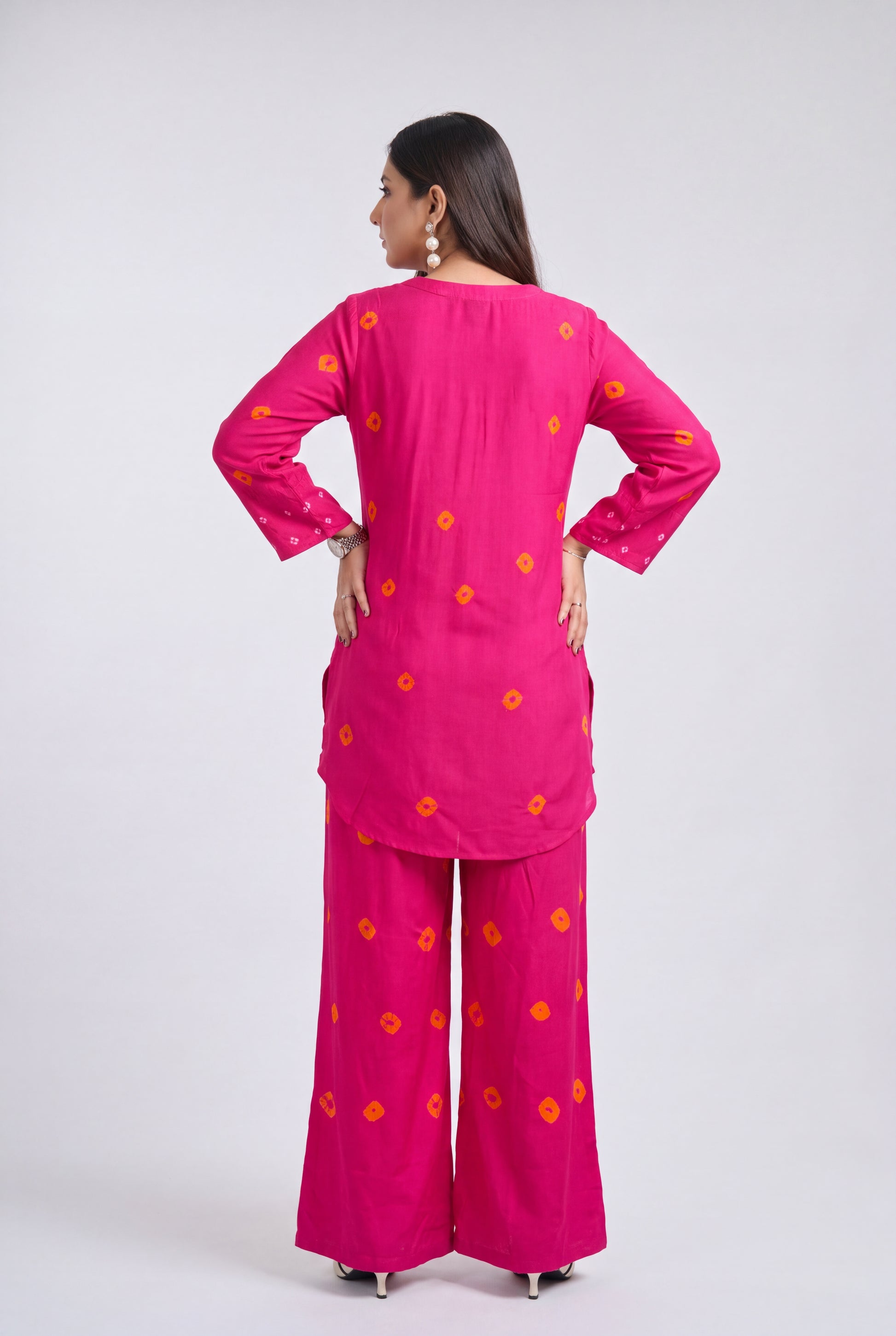 A woman is shown from the back wearing a bright pink two-piece kurta and trouser set with small orange floral motifs, set against a plain light gray studio background.