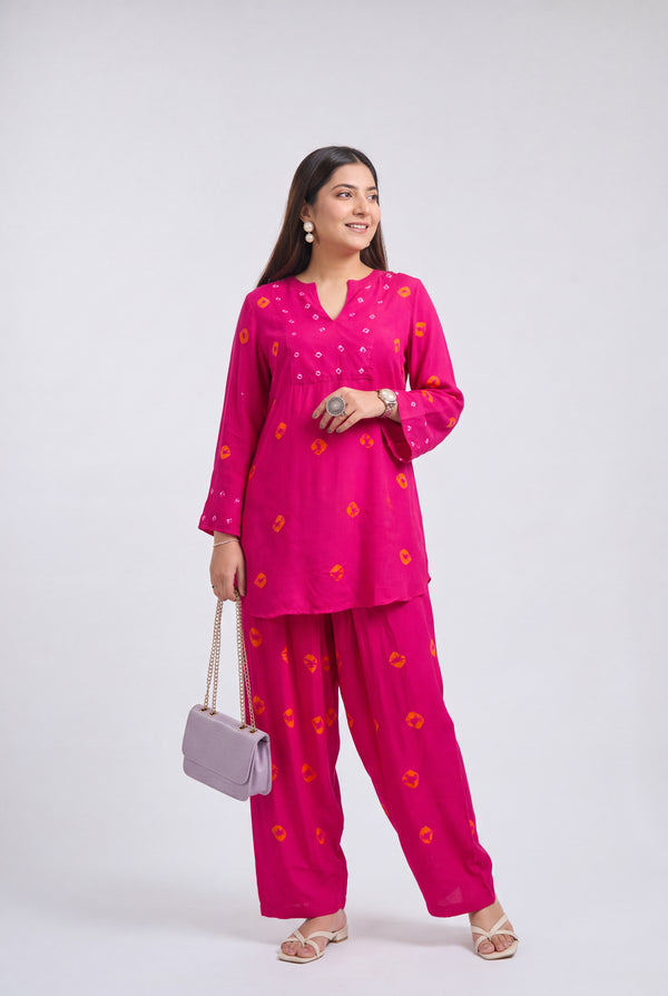 A woman modeling a bright pink two-piece kurta set with orange floral patterns, holding a light purple handbag against a plain light gray studio background.