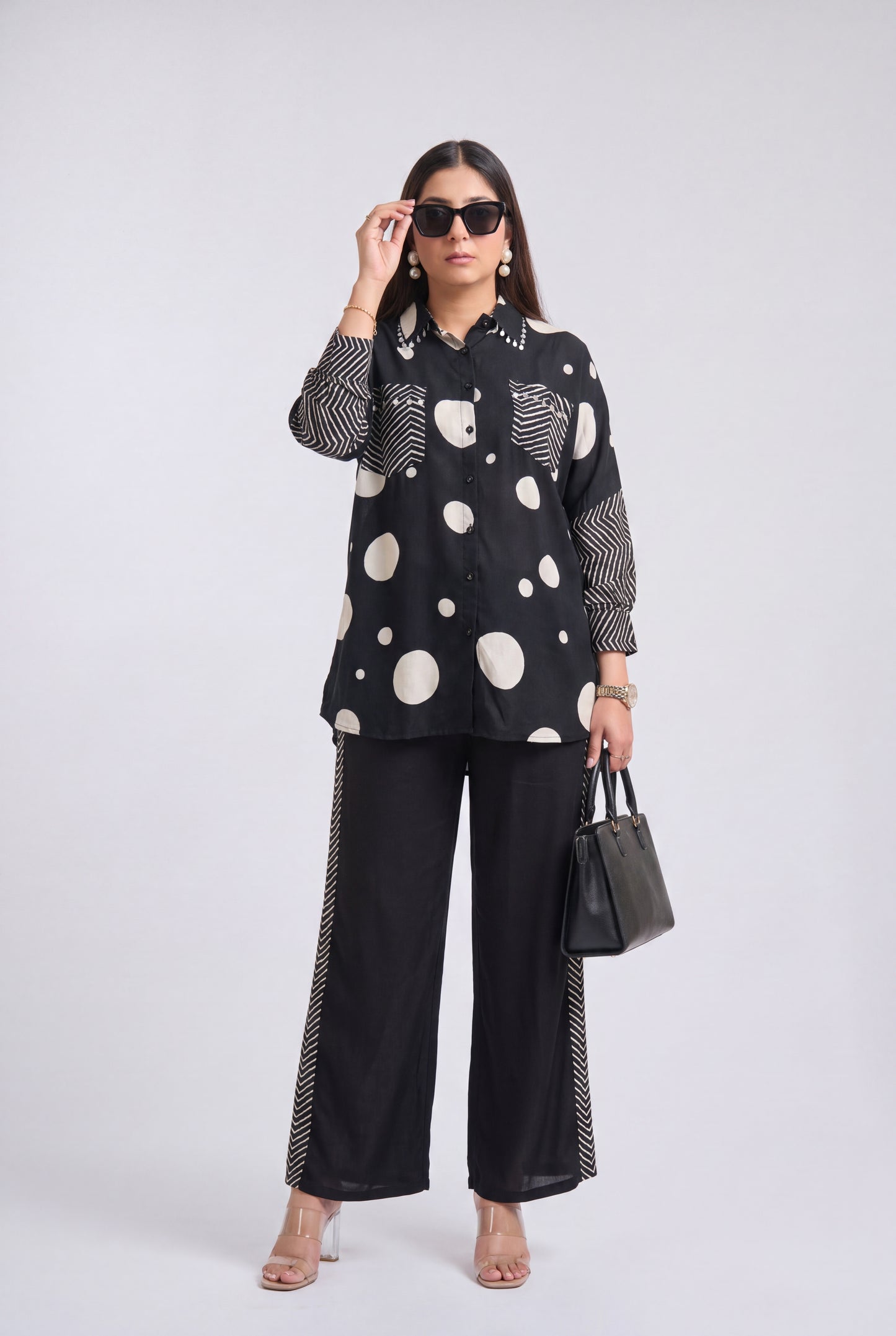 A woman modeling a black long-sleeve button-up shirt with large white polka dots and striped details, paired with black wide-leg pants featuring striped side panels, black sunglasses, and a black handbag, against a light gray studio background.