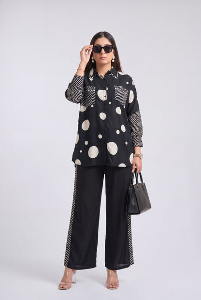 A woman modeling a black long-sleeve button-up shirt with large white polka dots and striped details, paired with black wide-leg pants featuring striped side panels, black sunglasses, and a black handbag, against a light gray studio background.