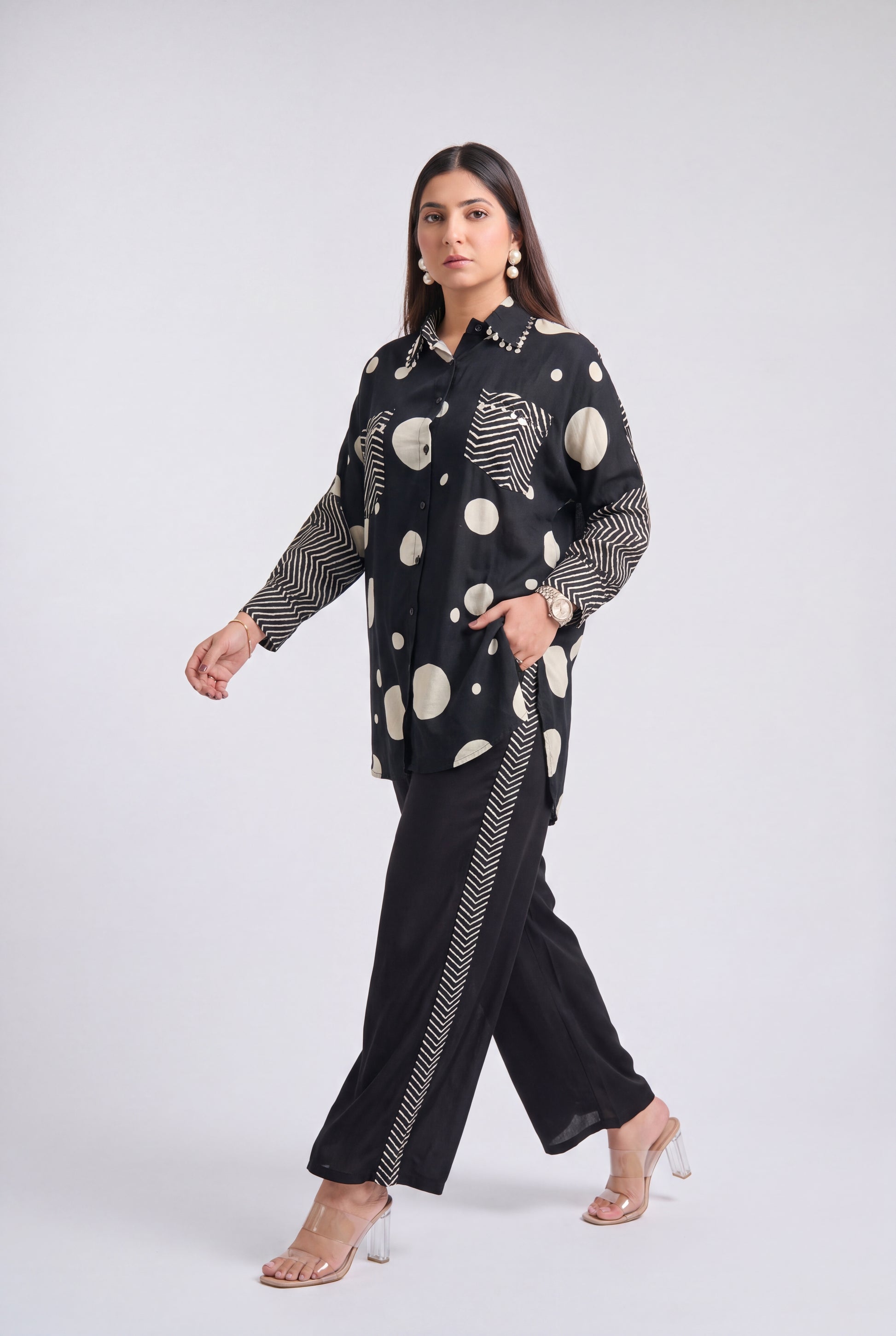 A woman models a black two-piece outfit featuring a long-sleeve button-up shirt with beige and white abstract circle patterns and matching wide-leg trousers with beige and white wavy stripe details along the side seams, set against a plain light gray studio background.
