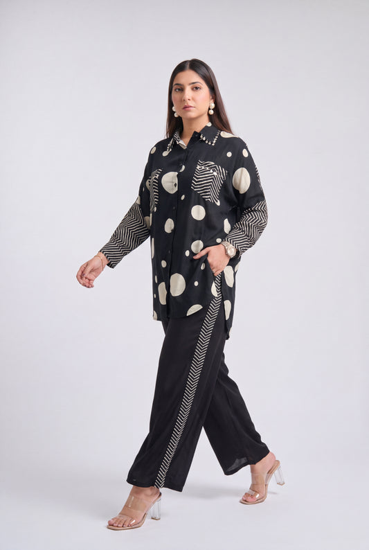 A woman models a black two-piece outfit featuring a long-sleeve button-up shirt with beige and white abstract circle patterns and matching wide-leg trousers with beige and white wavy stripe details along the side seams, set against a plain light gray studio background.