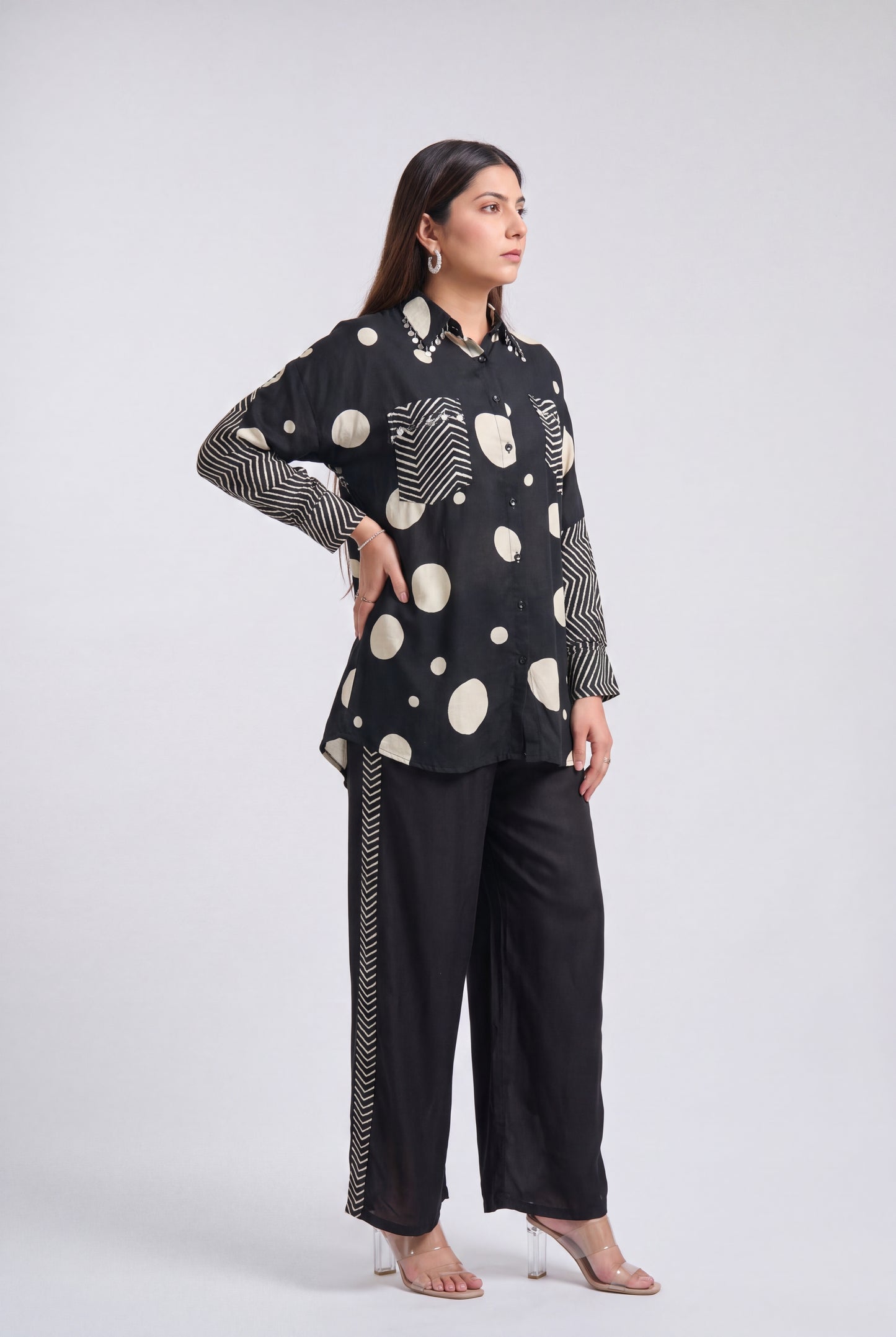 A woman modeling a black long-sleeve top with cream-colored polka dots, wavy line patterns, and striped cuffs, paired with black wide-leg trousers featuring a striped side detail, against a light gray studio background.