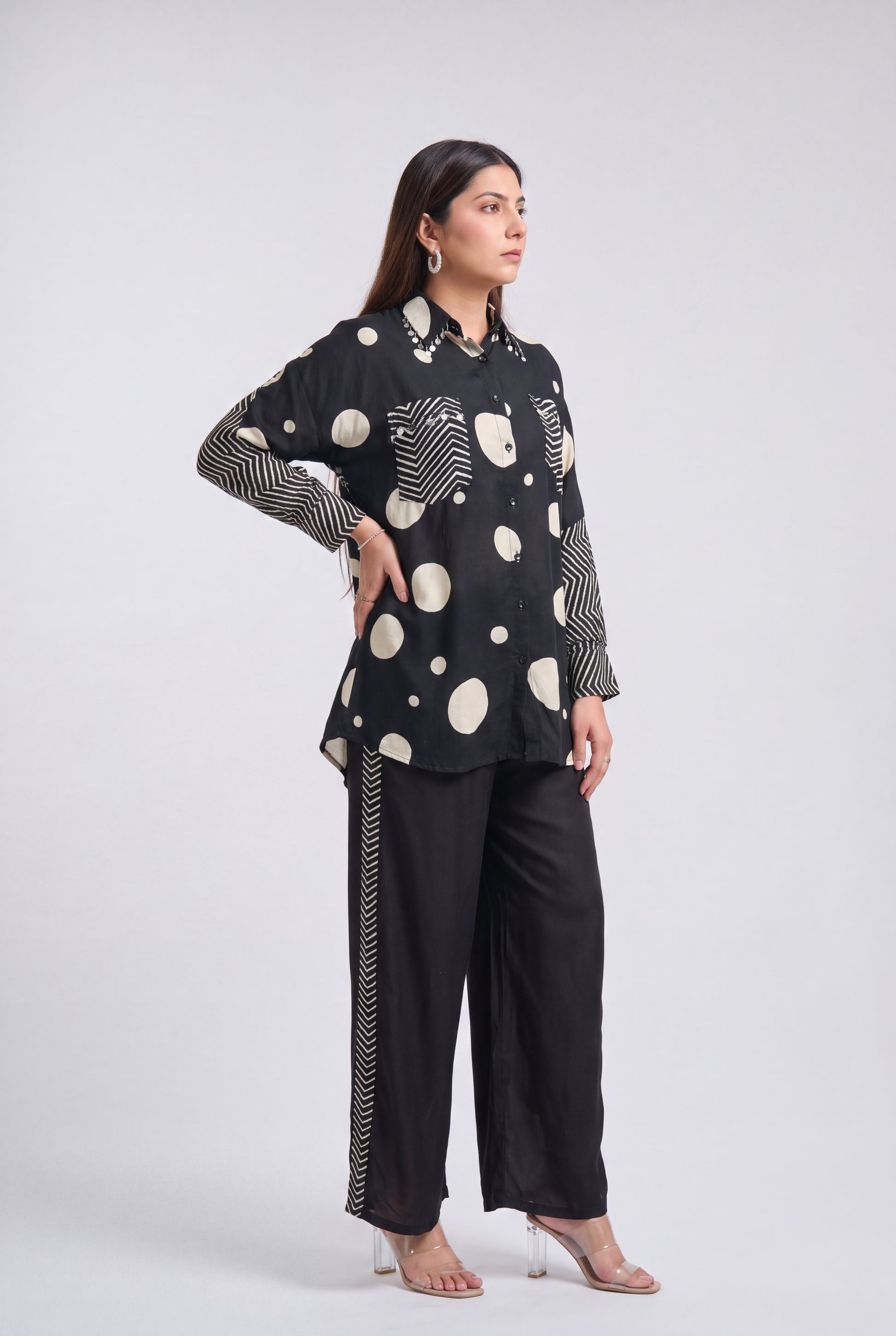 A woman modeling a black long-sleeve top with cream-colored polka dots, wavy line patterns, and striped cuffs, paired with black wide-leg trousers featuring a striped side detail, against a light gray studio background.