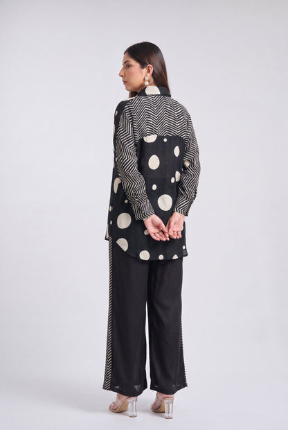 A woman is shown from the back wearing a black and white patterned kurta and wide-leg trousers set, standing against a light gray studio background.