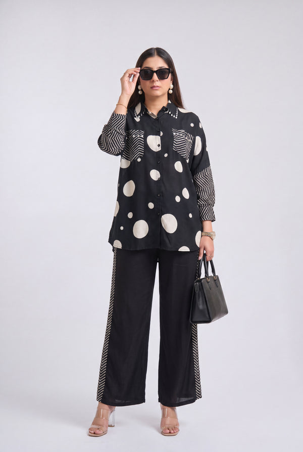 A woman modeling a black long-sleeve button-up shirt with large white polka dots and striped details, paired with black wide-leg pants featuring striped side panels, black sunglasses, and a black handbag, against a light gray studio background.