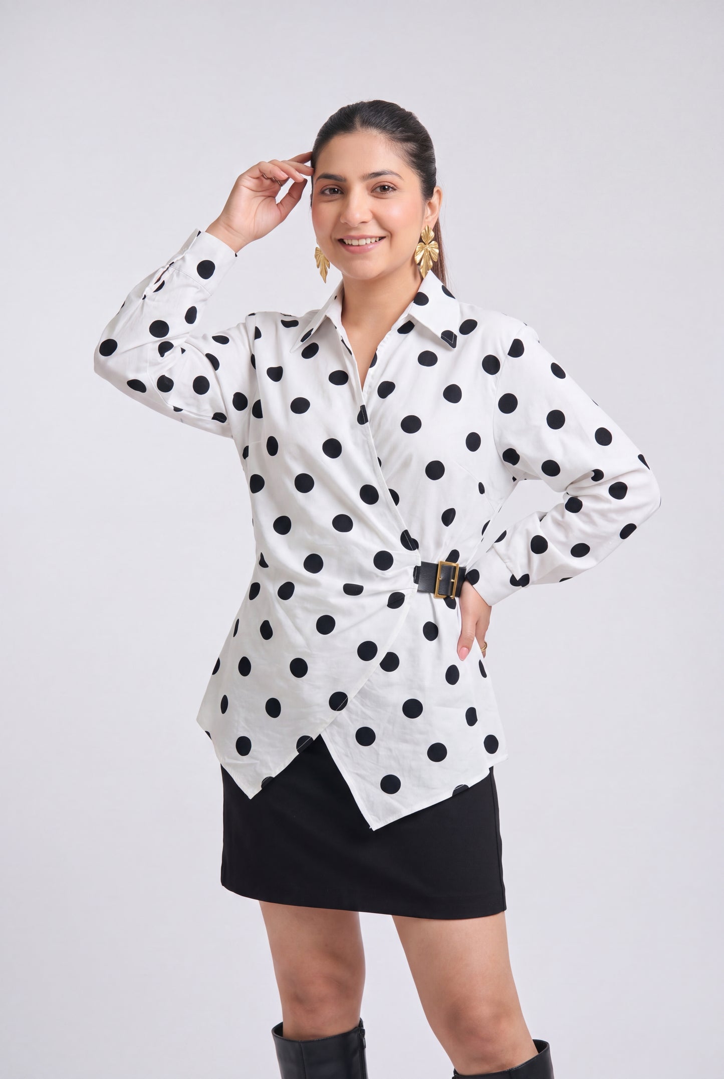 White and black polka dot wrap shirt with collared neckline, full sleeves, waist belt and asymmetrical hem, styled with a black mini skirt and knee-high boots.