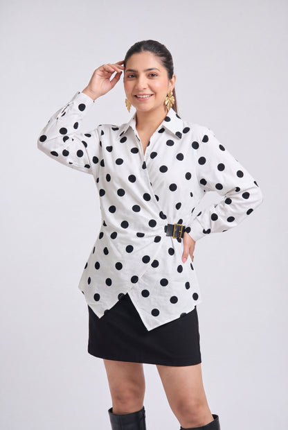 White and black polka dot wrap shirt with collared neckline, full sleeves, waist belt and asymmetrical hem, styled with a black mini skirt and knee-high boots.