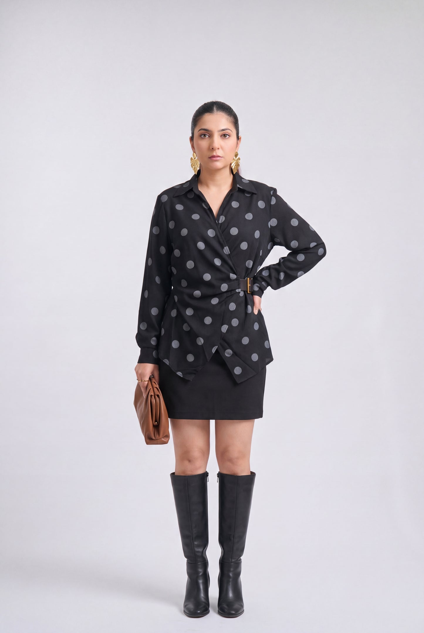 White and black polka dot wrap shirt with collared neckline, full sleeves, waist belt and asymmetrical hem, styled with a black mini skirt and knee-high boots.