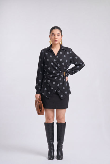 White and black polka dot wrap shirt with collared neckline, full sleeves, waist belt and asymmetrical hem, styled with a black mini skirt and knee-high boots.