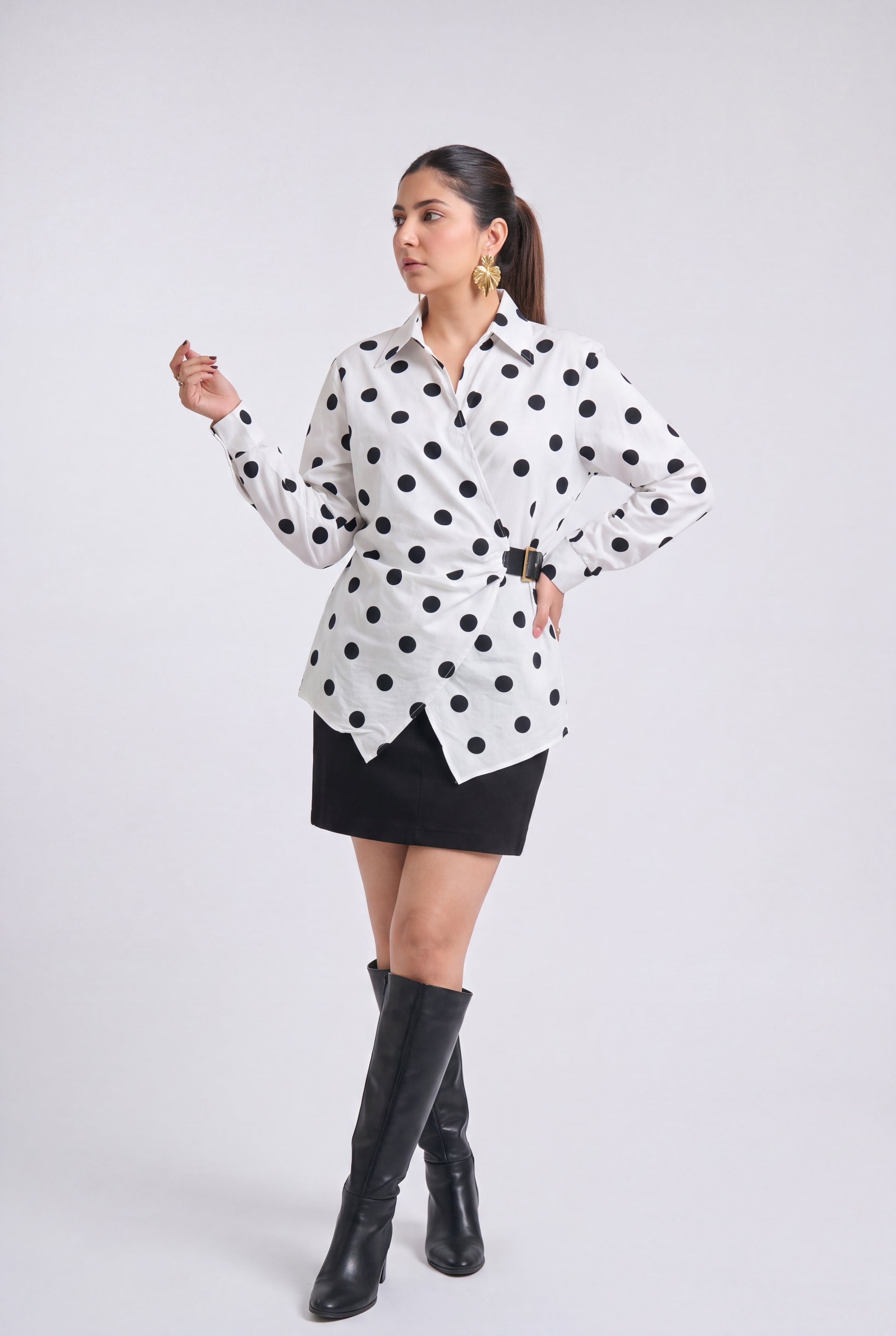 A woman modeling a white long-sleeve button-up shirt with black polka dots, styled with a black mini skirt, black knee-high boots, and gold hoop earrings against a light gray studio background.