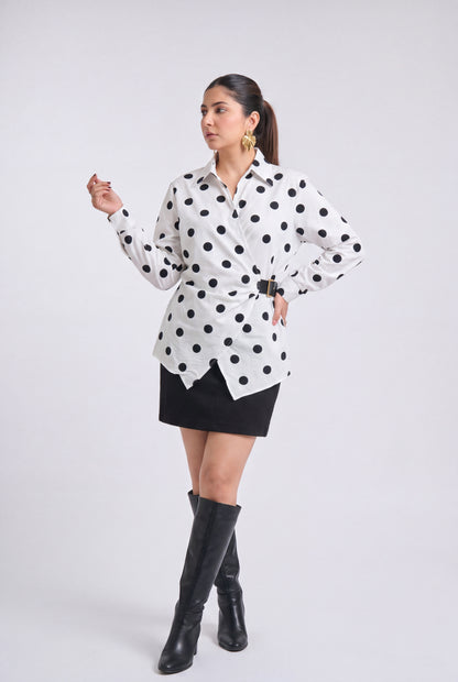 A woman modeling a white long-sleeve button-up shirt with black polka dots, styled with a black mini skirt, black knee-high boots, and gold hoop earrings against a light gray studio background.