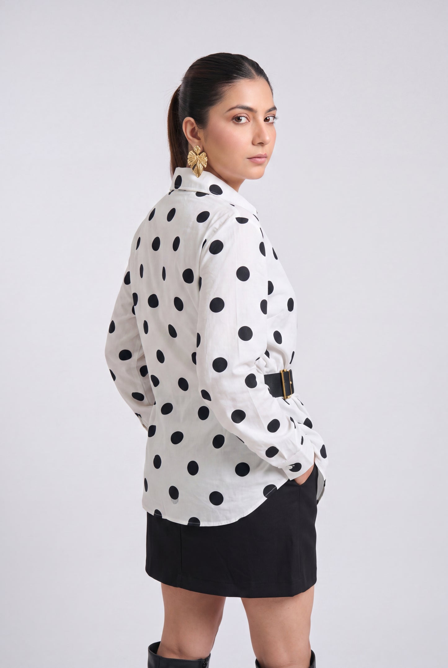 White and black polka dot wrap shirt with collared neckline, full sleeves, waist belt and asymmetrical hem, styled with a black mini skirt and knee-high boots.
