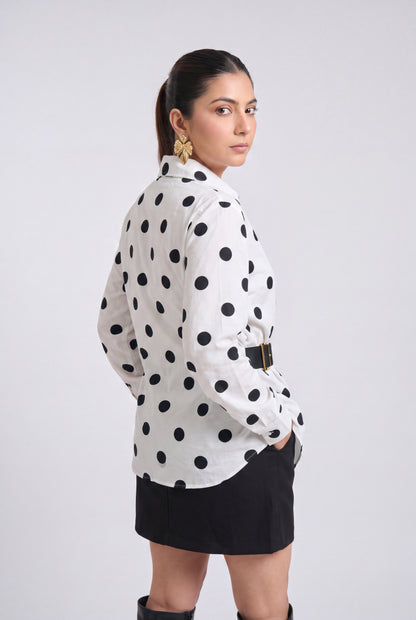White and black polka dot wrap shirt with collared neckline, full sleeves, waist belt and asymmetrical hem, styled with a black mini skirt and knee-high boots.