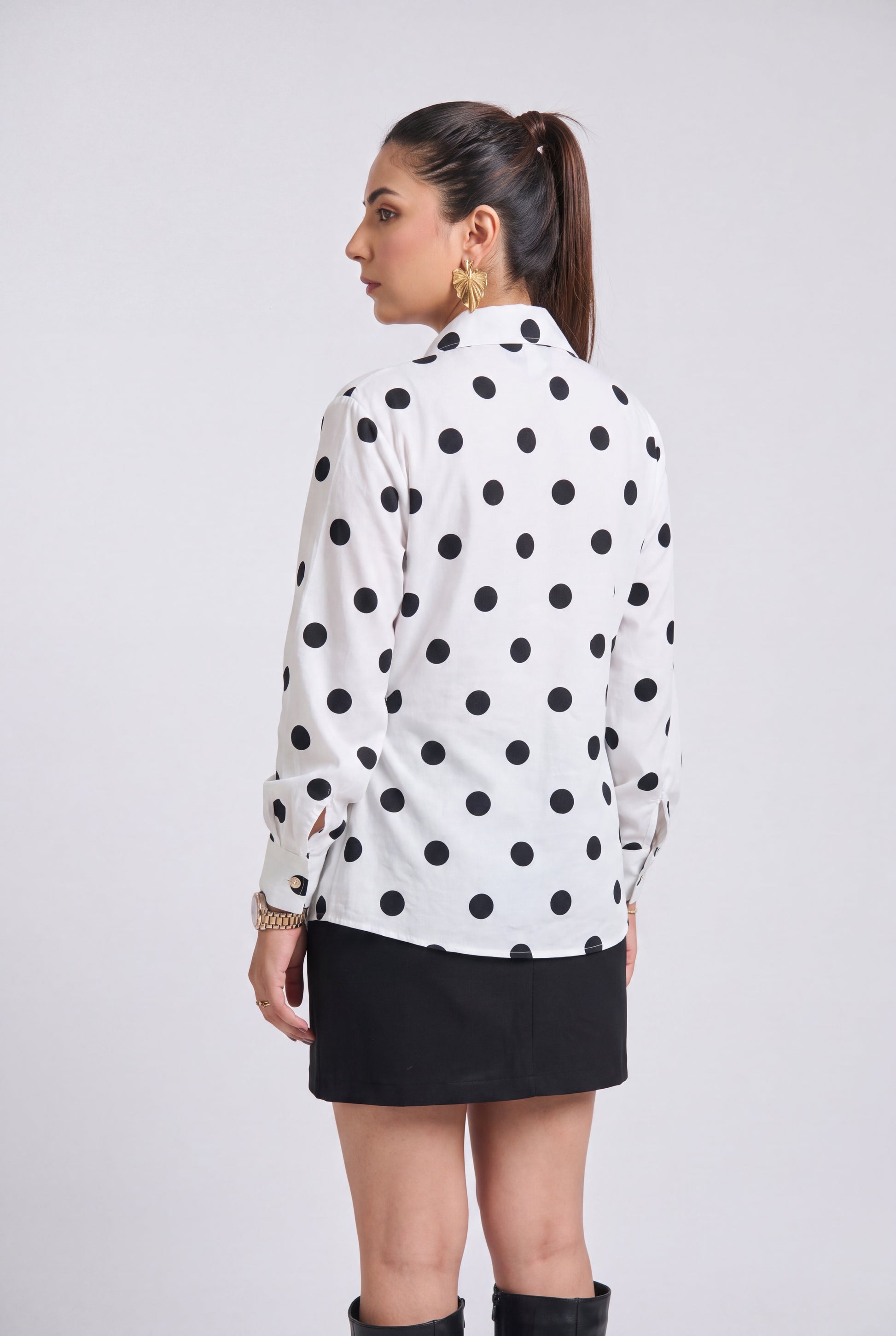 A woman shown from the back wearing a white long-sleeve button-up shirt with black polka dots, paired with a black mini skirt and black knee-high boots, against a light gray studio background.