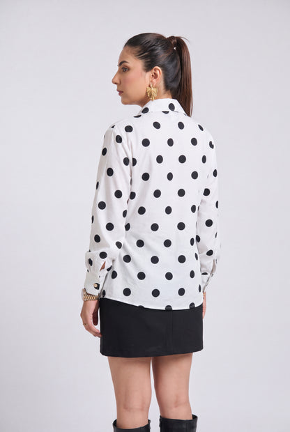 A woman shown from the back wearing a white long-sleeve button-up shirt with black polka dots, paired with a black mini skirt and black knee-high boots, against a light gray studio background.
