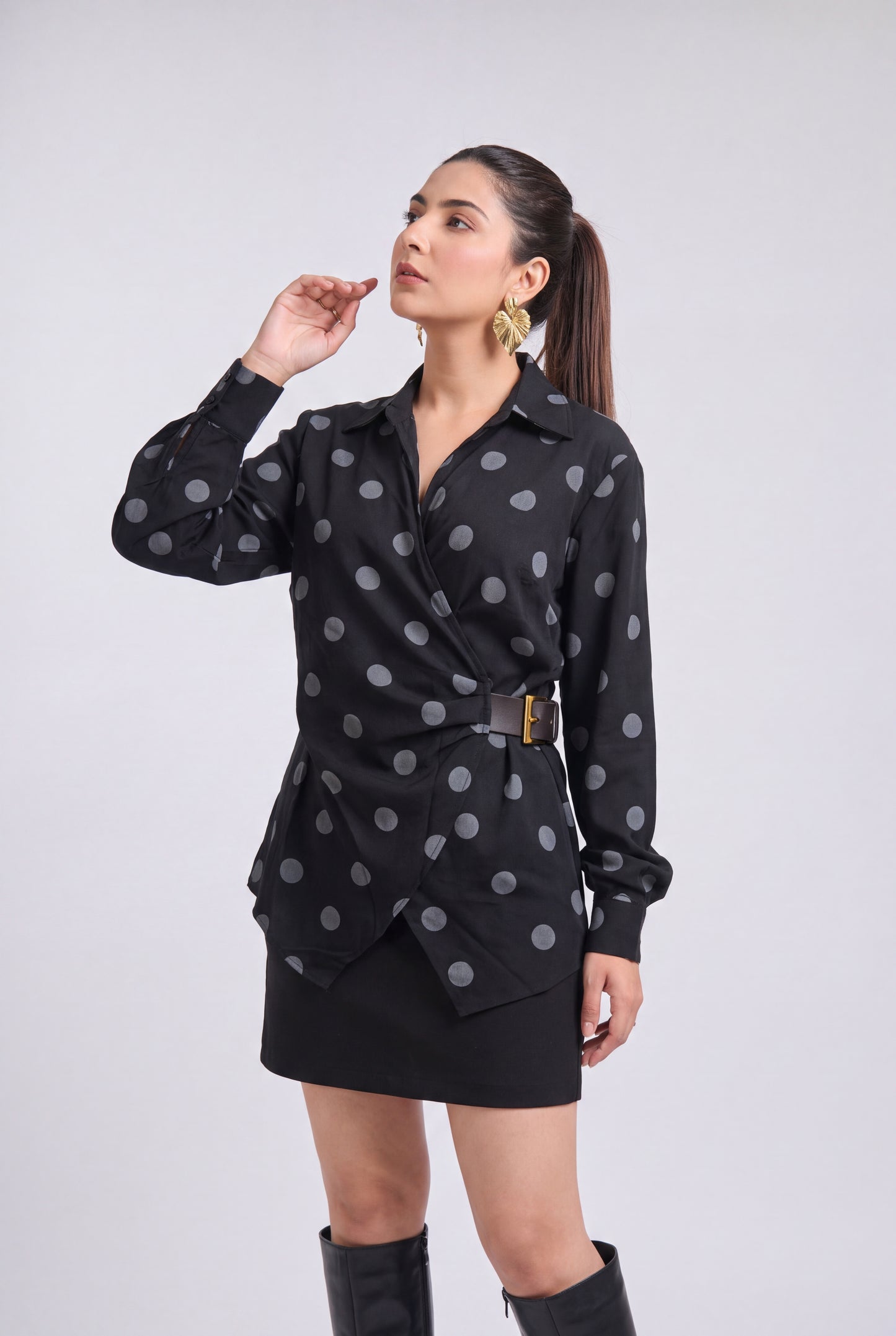 A woman modeling a black long-sleeve wrap-style blouse with gray polka dots and a matching black mini skirt, paired with black knee-high boots, against a light gray studio background.